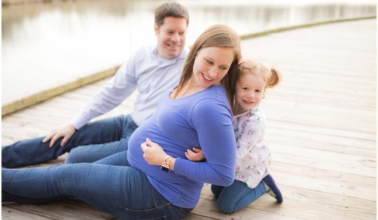 Family of three sitting on the wooden dock with little girl holding mom's pregnant belly.